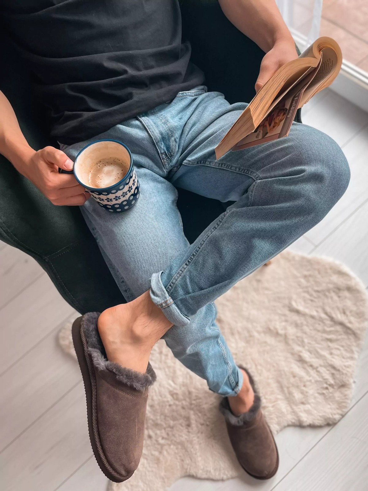 Brązowe kapcie z ciemnym wnętrzem na stopach osoby z książką i kubkiem